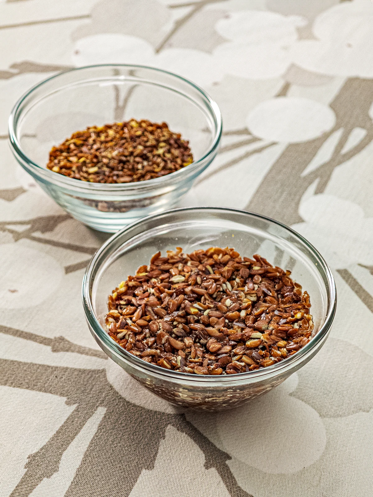 Two glass bowls are placed on a tablecloth with a branched pattern. Both bowls contain ground flaxseed. In the front glass bowl, the flaxseed is completely swollen.