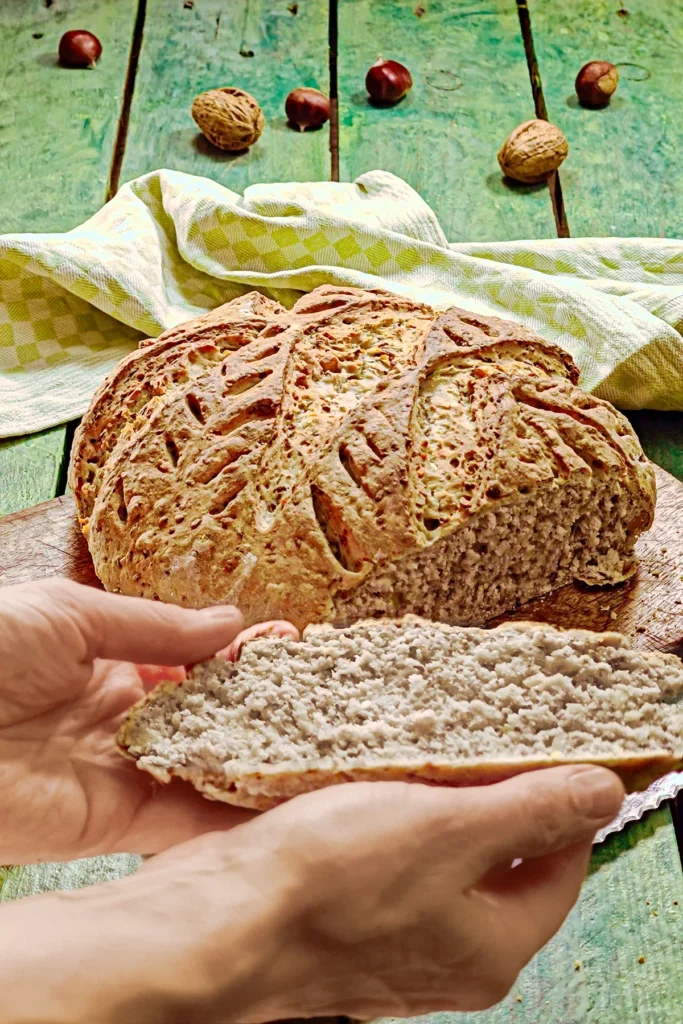 A wooden cutting board with a chestnut bread lies on a green wooden table. Two hands present a slice of chestnut bread. A green kitchen towel, chestnuts and walnuts lie around in the background.