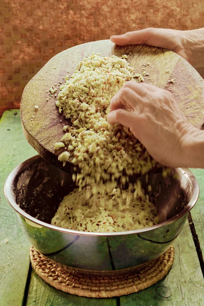 Two hands add the chopped chestnuts from a wooden board to the dough in the stainless steel bowl with the mixed bread flour mixture. The bowl stands on a raffia coaster on a green wooden table.