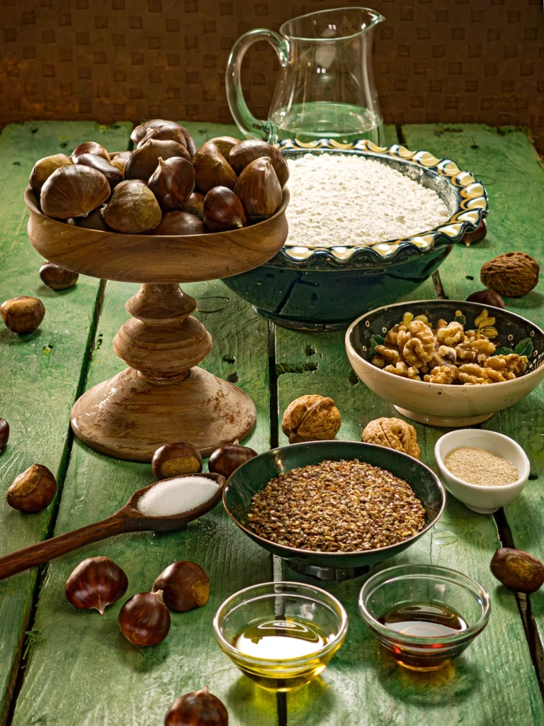 There are various bowls on a green wooden table. A large one in the background with spelt flour, in front of it there are bowls with walnuts, linseed, dry yeast, olive oil and maple syrup. Also there is an wooden etagere with fresh chestnuts, in front of it a wooden spoon with salt. Behind the bowl of flour is a glass jug of water. A few chestnuts and walnuts are scattered around.