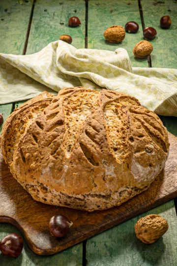 A wooden chopping board with a whole chestnut loaf lies on a green wooden table. Behind it is a green kitchen towel. A few chestnuts and walnuts are scattered around it.