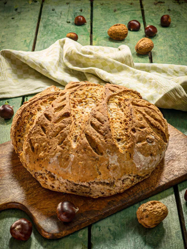 A wooden chopping board with a whole chestnut loaf lies on a green wooden table. Behind it is a green kitchen towel. A few chestnuts and walnuts are scattered around it.