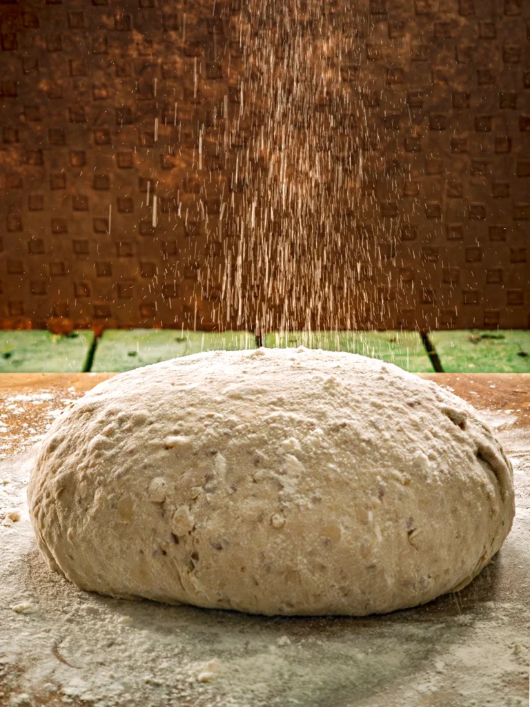 Flour trickles from above onto the dough for a chestnut loaf. The ball of dough lies on a wooden cutting board.