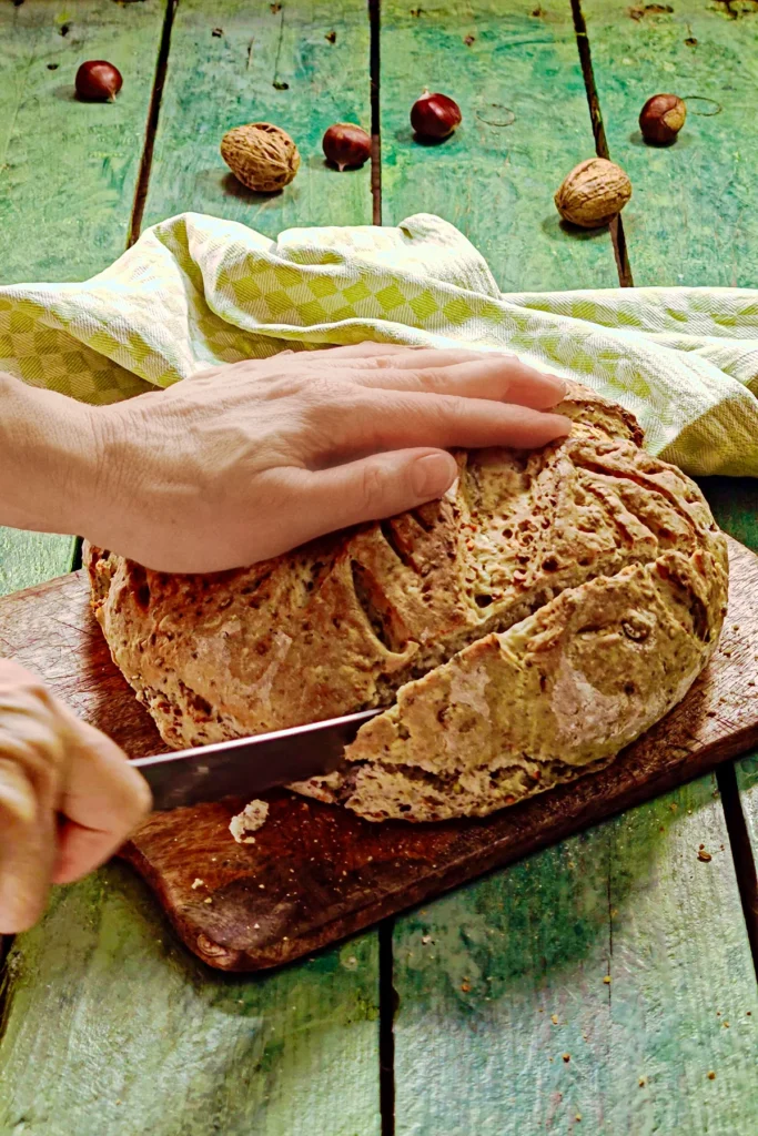 A wooden chopping board with a chestnut loaf lies on a green wooden table. One hand holds the bread, another hand with a knife begins to cut off the first slice of bread. 