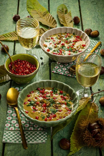 Dinner is served, two soup bowls with chestnut soup and topping stand on a green wooden table surface. Both soup bowls have next to them a green patterned napkin with a golden spoon on top and a glass of white alkohlfree wine. Between the bowls is a small green bowl with pomegranate seeds and a golden teaspoon. On the green base are scattered chestnut leaves and whole chestnuts in shells.