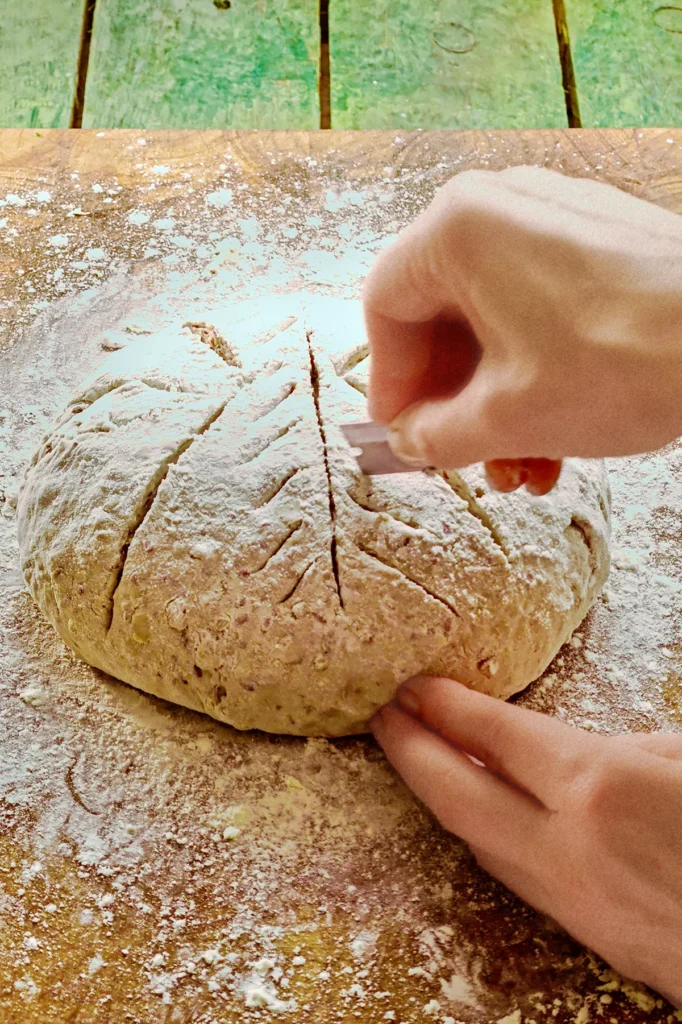 Two hands decorate the ball of dough, the right hand holds a razor blade that carves a pattern into the surface of the dough. The chestnut bread dough lies on a wooden cutting board.