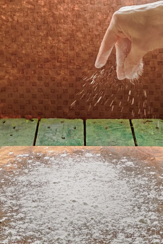 One hand sprinkles flour on the wooden cutting board.