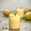 Two servings of vegan mango lassi with mangos in the background and fresh mint as a garnish