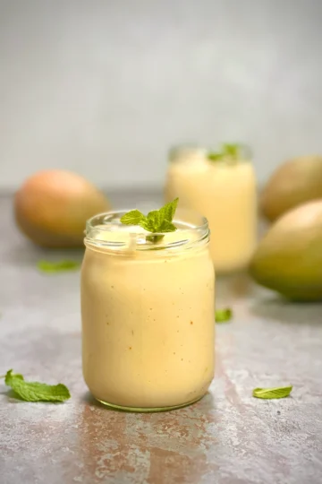 Two servings of vegan mango lassi with mangos in the background and fresh mint as a garnish