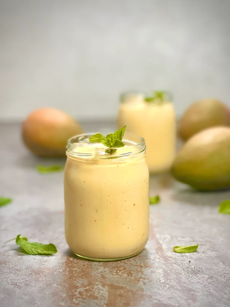 Two servings of vegan mango lassi with mangos in the background and fresh mint as a garnish