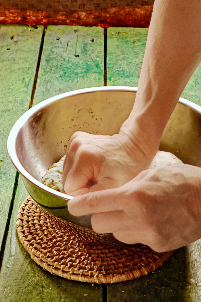 A hand kneads the dough in the stainless steel bowl. The bowl stands on a raffia coaster on a green wooden table.