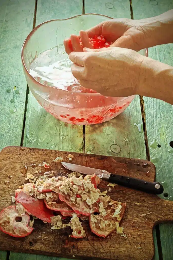 A wooden cutting board with a kitchen knife is filled with the remains of the peel from the pomegranate seeds removing process. Behind there is a large glass bowl filled with water in which pomegranate seeds lie at the bottom. Two hands are over the bowl, one hand has pomegranate seeds in the palm. The base where the wooden kitchen board and the glass bowl stands on, is made of green-painted wooden planks.