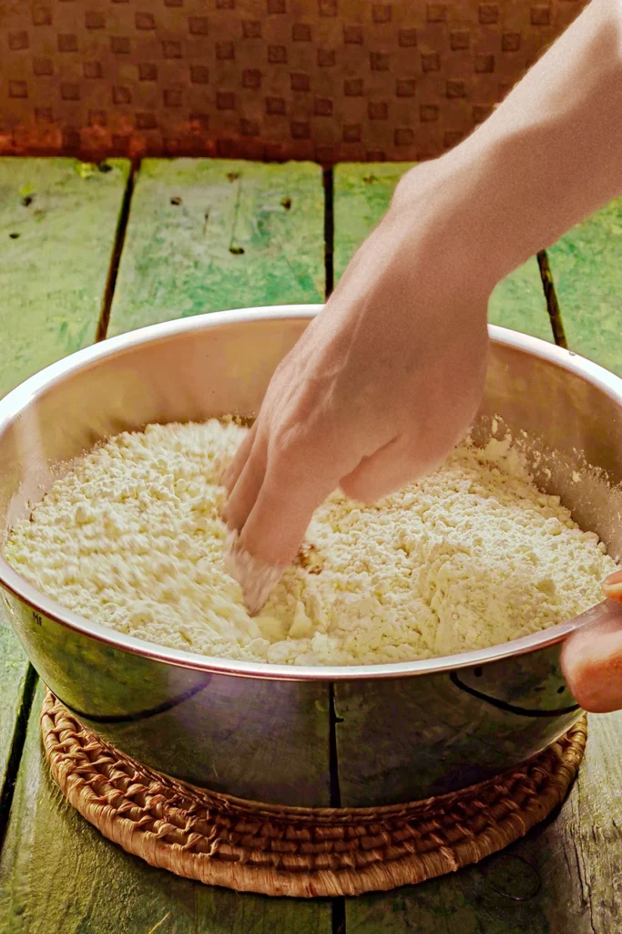 Dip a hand into the stainless steel bowl with the bread flour mixture to mix the ingredients. The bowl stands on a raffia coaster on a green wooden table.