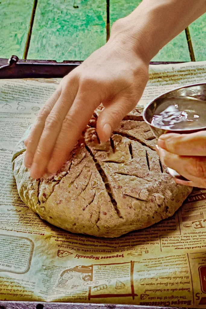 One hand carefully taps a little water onto the floured surface of the dough. The chestnut bread dough is placed on a wooden cutting board.