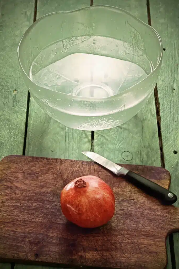 A pomegranate lies on a wooden kitchen board, with a kitchen knife next to it. In the background is a large glass bowl filled with water. The base is made of green-painted wooden planks.