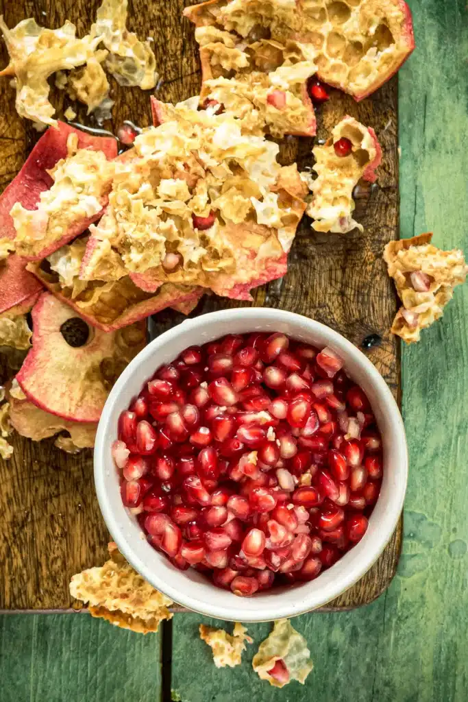 The top view shows a wooden cutting board lying on a green wooden base, filled with the remains of the peel from the pomegranate coring process. In front of the board is a white bowl filled with pomegranate seeds.