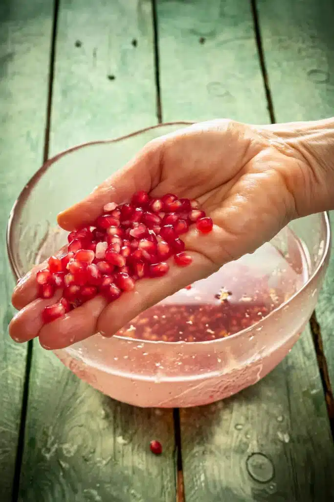 One hand shows pomegranate seeds in the palm. Behind it is a large glass bowl filled with water, in which pomegranate seeds lie at the bottom, the water somewhat cloudy from the coring.