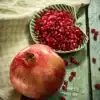 A pomegranate lies on a green wooden base. Behind it is a flat green leaf-shaped bowl filled with pomegranate seeds. A few seeds are scattered on the green base. Next to it is a green kitchen towel.