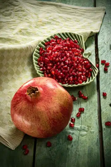 A pomegranate lies on a green wooden base. Behind it is a flat green leaf-shaped bowl filled with pomegranate seeds. A few seeds are scattered on the green base. Next to it is a green kitchen towel.