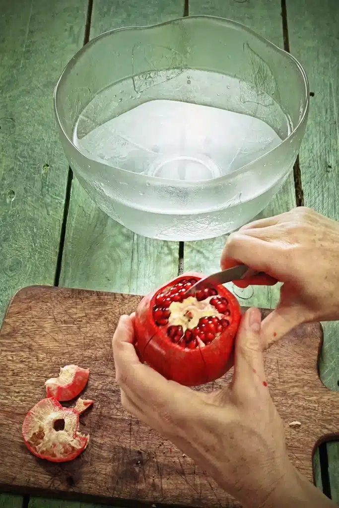 Two hands are holding a pomegranate with the skin missing at the top in the form of a lid. In one hand is a kitchen knife that has carved the skin of the pomegranate lengthwise. Below the pomegranate is a wooden kitchen board on a green wooden base. In the background is a large glass bowl filled with water.