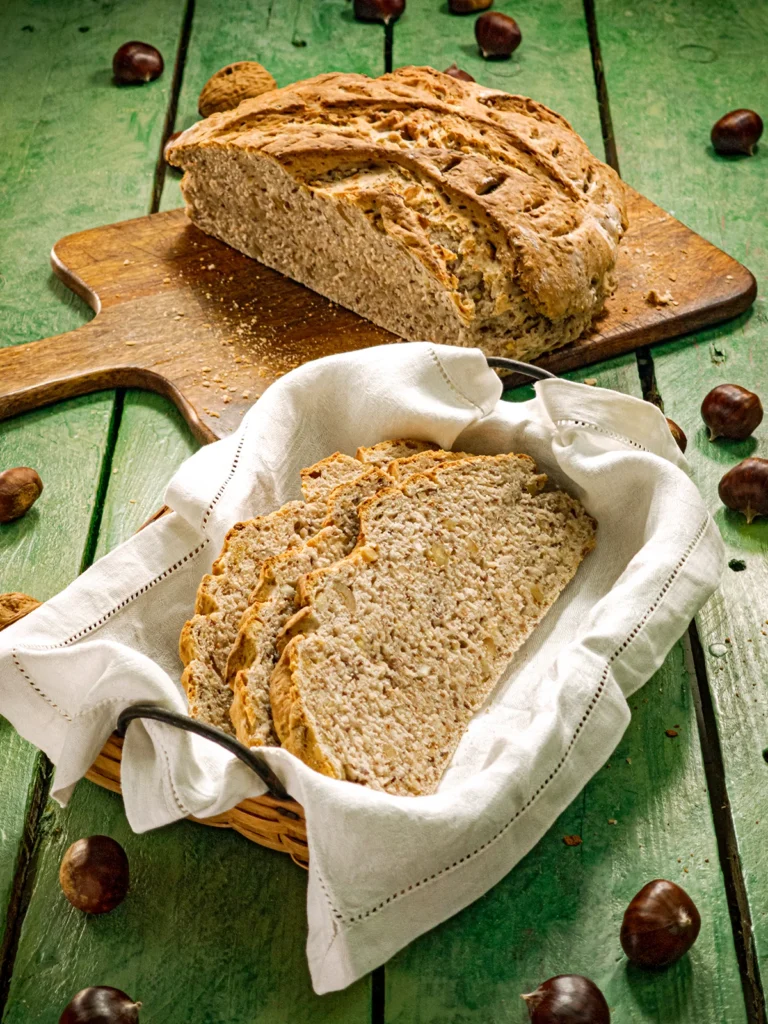 A wooden chopping board with a sliced chestnut bread lies on a green wooden table. In front of it is a bread basket with a white linen cover and a few slices of chestnut bread. A few chestnuts and walnuts are scattered around.