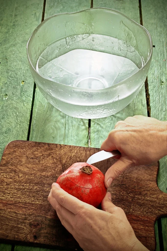 Two hands hold a pomegranate. In one hand is a kitchen knife that has carved a circle around the blossom of the fruit. The pomegranate lies on a wooden kitchen board on a green wooden base. In the background is a large glass bowl filled with water.