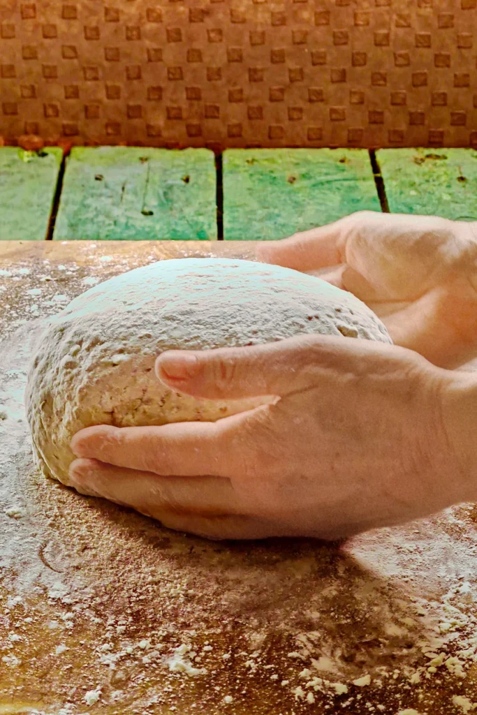 Two hands hold the ball of dough and shape it carefully. The chestnut bread dough is placed on a wooden cutting board.