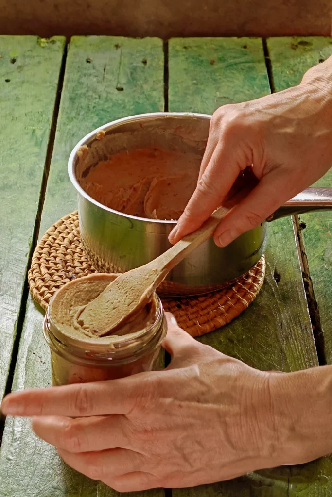 A raffia trivet with a stainless steel pot lies on a green wooden table. The pot contains the pureed chestnut spread. In the foreground, one hand holds a jar filled with chestnut spread. The other hand holds a wooden spatula and presses the cream into the jar.