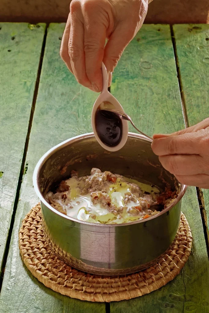 A raffia trivet with a stainless steel pot lies on a green wooden table. In the pot is the ready-cooked mixture of vegetables, on top is vegan cream. One hand holds a white bowl  with brown sugar molasse and add it to the pot with a spoon.