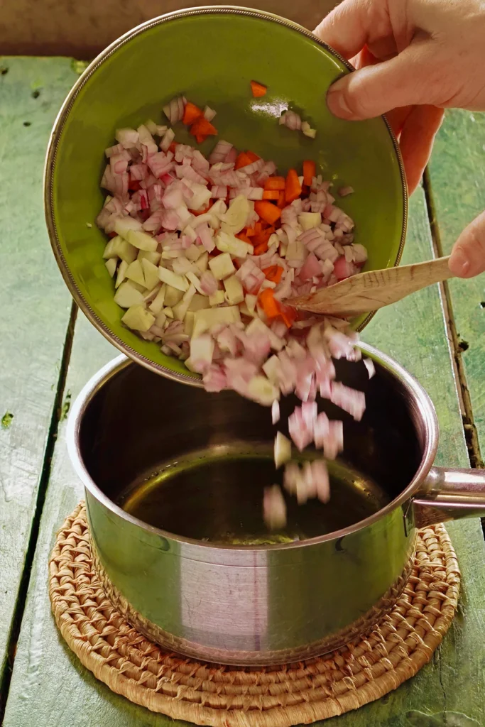 A raffia trivet with a stainless steel pot lies on a green wooden table. In the pot is the chopped mixture of vegetables, a hand holds a green bowl over the pot, the bowl is filled with chopped vegetables. One hand holds a wooden spatula and lifts the chestnuts into the pot.