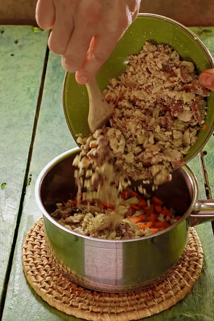 A raffia trivet with a stainless steel pot lies on a green wooden table. In the pot is the chopped mixture of vegetables, a hand holds a green bowl over the pot, the bowl is filled with chopped chestnuts. One hand holds a wooden spatula and lifts the chestnuts into the pot.