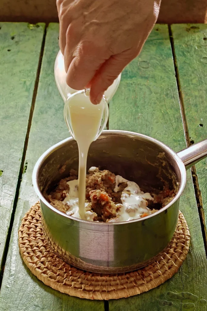 A raffia trivet with a stainless steel pot lies on a green wooden table. In the pot is the ready -cooked mixture of vegetables, on top is vegan cream. One hand holds a jar with vegan cream and add it to the pot.