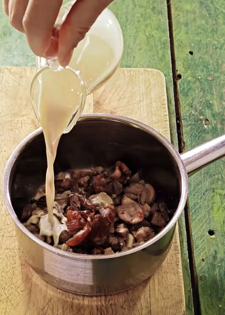 A wooden cutting board lies on a green wooden table with a stainless steel pot on top. In the pot is the mixture of ingredients for créme de marrons. One hand add roat milk to the pot.