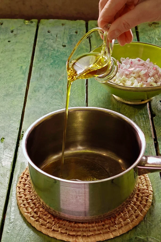 A raffia trivet with a stainless steel pot lies on a green wooden table. One hand holds a jar with olive oil and add it to the pot. In the backgrouns is a green bowl with chopped vegetables.