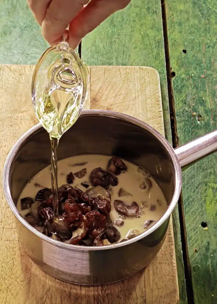 A wooden cutting board lies on a green wooden table with a stainless steel pot on top. In the pot is the mixture of ingredients for créme de marrons. One hand add rapeseed oil to the pot.