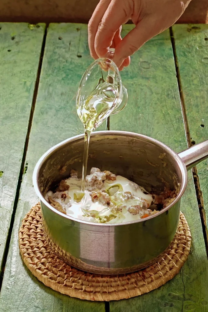 A raffia trivet with a stainless steel pot lies on a green wooden table. In the pot is the ready -cooked mixture of vegetables, on top is vegan cream. One hand holds a jar with rapeseed oil and add it to the pot.