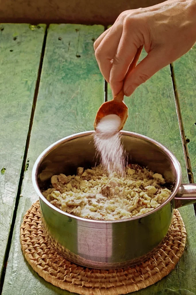 A raffia trivet with a stainless steel pot lies on a green wooden table. In the pot are the chopped mixture of vegetables, on top are the chestnuts. One hand holds a roughly carved spoon that pours salt over the chestnuts.