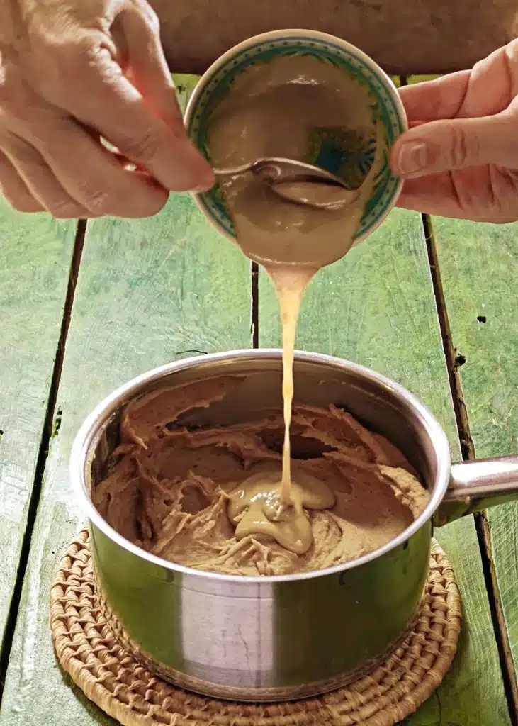 A raffia trivet with a stainless steel pot lies on a green wooden table. In the pot is the ready-cooked mixture of vegetables, on top is vegan cream. One hand holds a small green bowl with tahini and add it to the pot with a spoon.