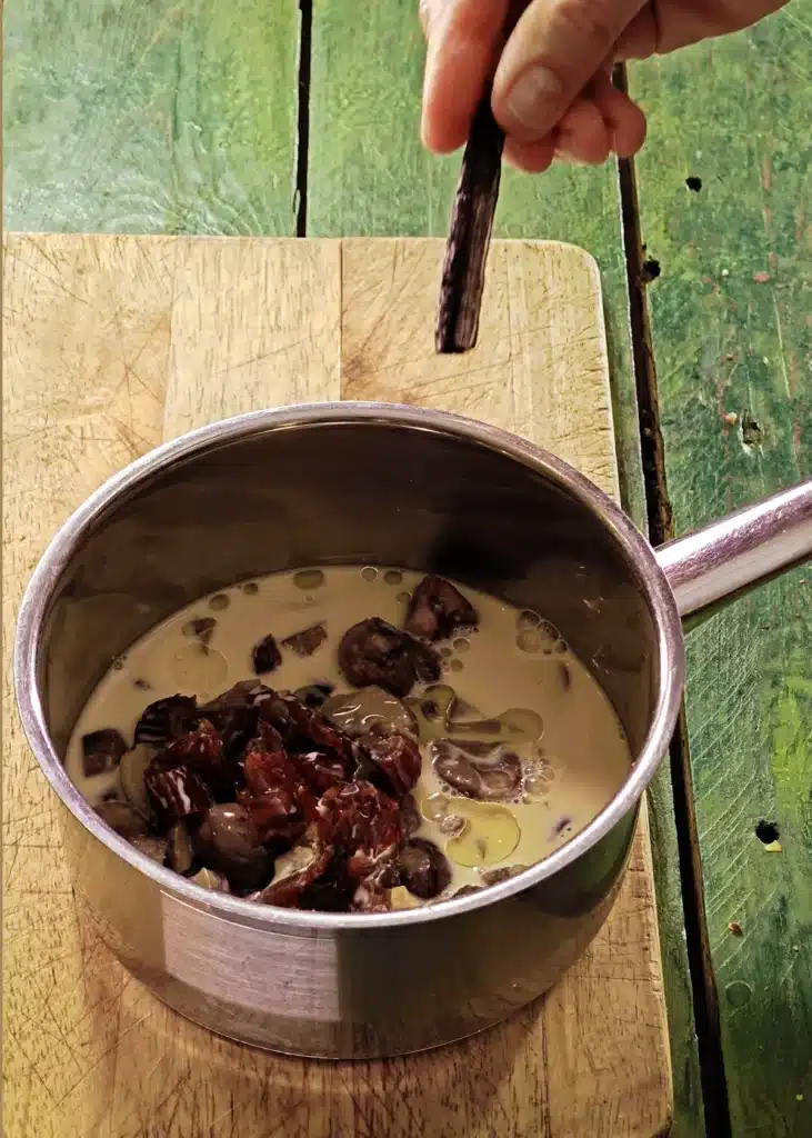 A wooden cutting board lies on a green wooden table with a stainless steel pot on top. In the pot is the mixture of ingredients for créme de marrons. One hand add vanilla pod to the pot.