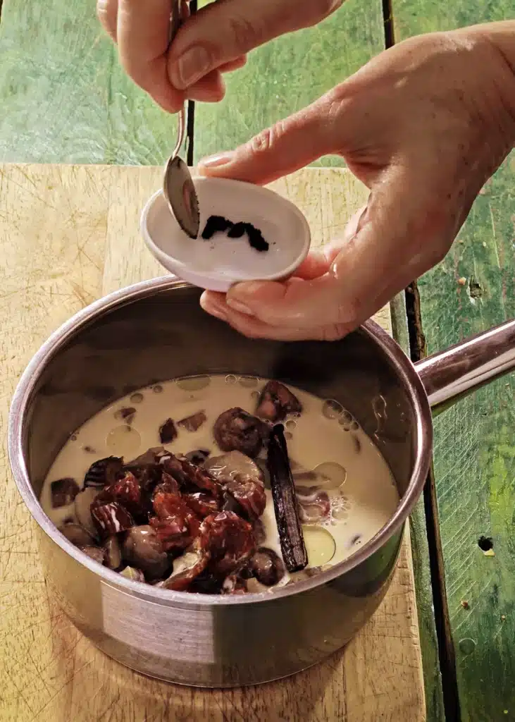 A wooden cutting board lies on a green wooden table with a stainless steel pot on top. In the pot is the mixture of ingredients for créme de marrons. One hand holds a small white bowl of fresh vanilla. A spoon in the other hand adds the vanilla to the pot.