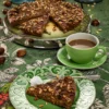 A slice of traditional Italian Castagnaccio on a decorative green plate, topped with pine nuts and rosemary, served with a cup of coffee on a vibrant green tablecloth adorned with chestnuts and walnuts.