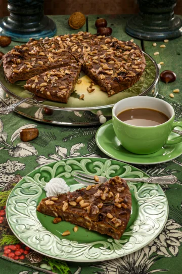 A slice of traditional Italian Castagnaccio on a decorative green plate, topped with pine nuts and rosemary, served with a cup of coffee on a vibrant green tablecloth adorned with chestnuts and walnuts.