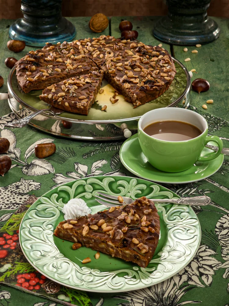 A slice of traditional Italian Castagnaccio on a decorative green plate, topped with pine nuts and rosemary, served with a cup of coffee on a vibrant green tablecloth adorned with chestnuts and walnuts.