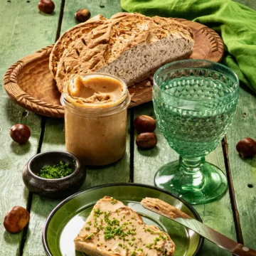 The meal is served on a green wooden table: a loaf of bread with chestnut spread, sprinkled with fresh chopped chives. The bread lies on a green plate, next to it a used wooden knife. Behind is a green glass with water, a preserving jar with the chestnut spread, and a flat black bowl with the chopped chives; further in the background is a flat wooden bowl with a sliced loaf of bread and a green linen kitchen towel. Scattered around everything are fresh chestnuts in shells.