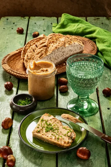 The meal is served on a green wooden table: a loaf of bread with chestnut spread, sprinkled with fresh chopped chives. The bread lies on a green plate, next to it a used wooden knife. Behind is a green glass with water, a preserving jar with the chestnut spread, and a flat black bowl with the chopped chives; further in the background is a flat wooden bowl with a sliced loaf of bread and a green linen kitchen towel. Scattered around everything are fresh chestnuts in shells.