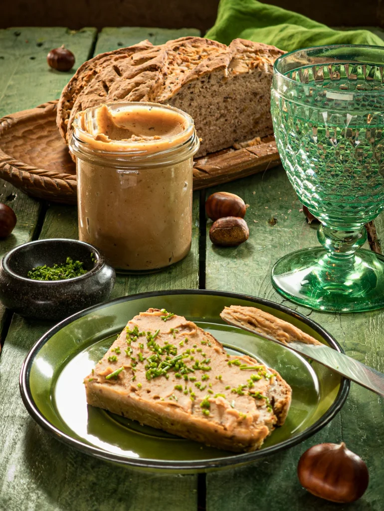 The meal is served on a green wooden table: a loaf of bread with chestnut spread, sprinkled with fresh chopped chives. The bread lies on a green plate, next to it a used wooden knife. Behind is a green glass with water, a preserving jar with the chestnut spread, and a flat black bowl with the chopped chives; further in the background is a flat wooden bowl with a sliced loaf of bread and a green linen kitchen towel. Scattered around everything are fresh chestnuts in shells.