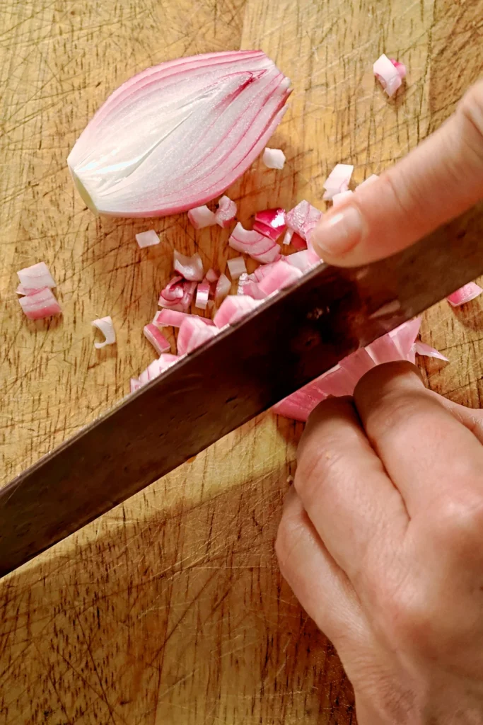 Chopped shallots lie on a wooden board, two hands hold a large knife and cut the shallots into small pieces.