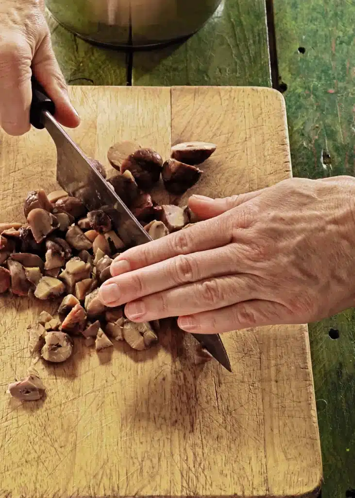 A wooden cutting board lies on a green wooden table with dates. Hands are holding a huge knife and chop the chestnuts.