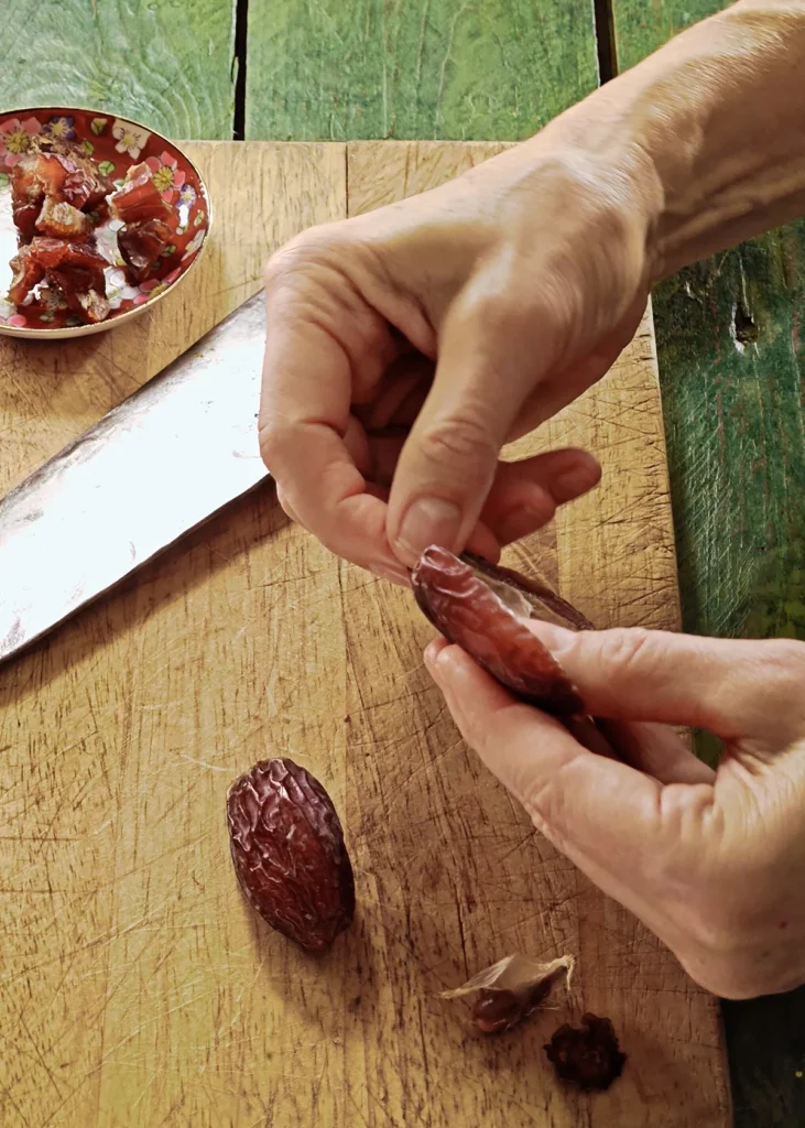 A wooden cutting board lies on a green wooden table with dates. Hands are removing the pit and chopping the date. In the background is a small bowl of chopped dates.