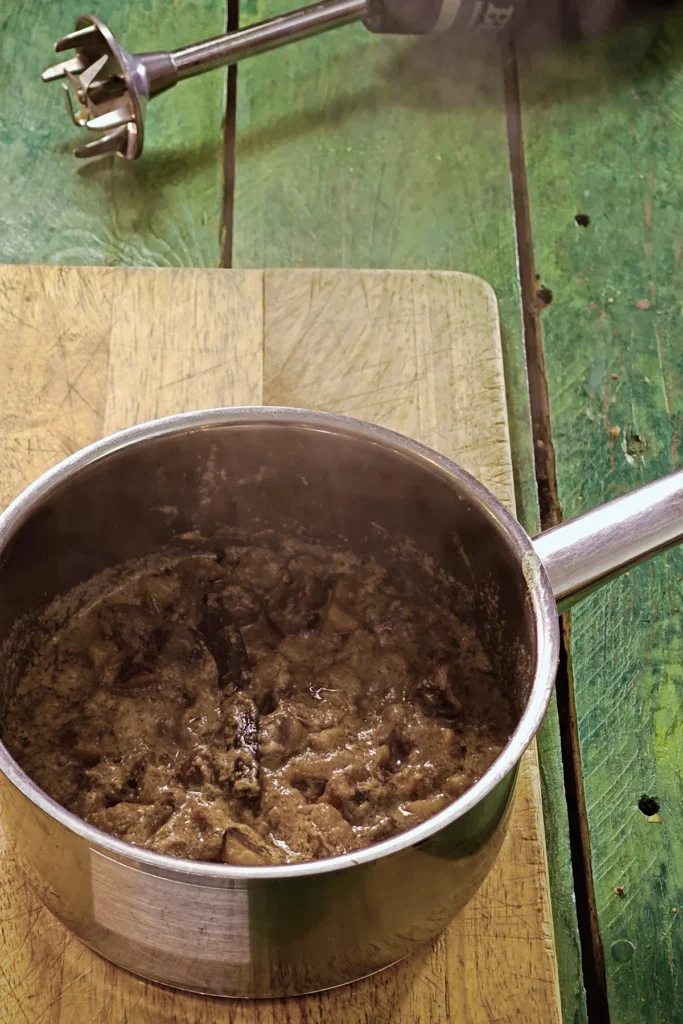 A wooden cutting board lies on a green wooden table with a stainless steel pot on top. In the pot is the freshly cooked mixture for créme de marrons.